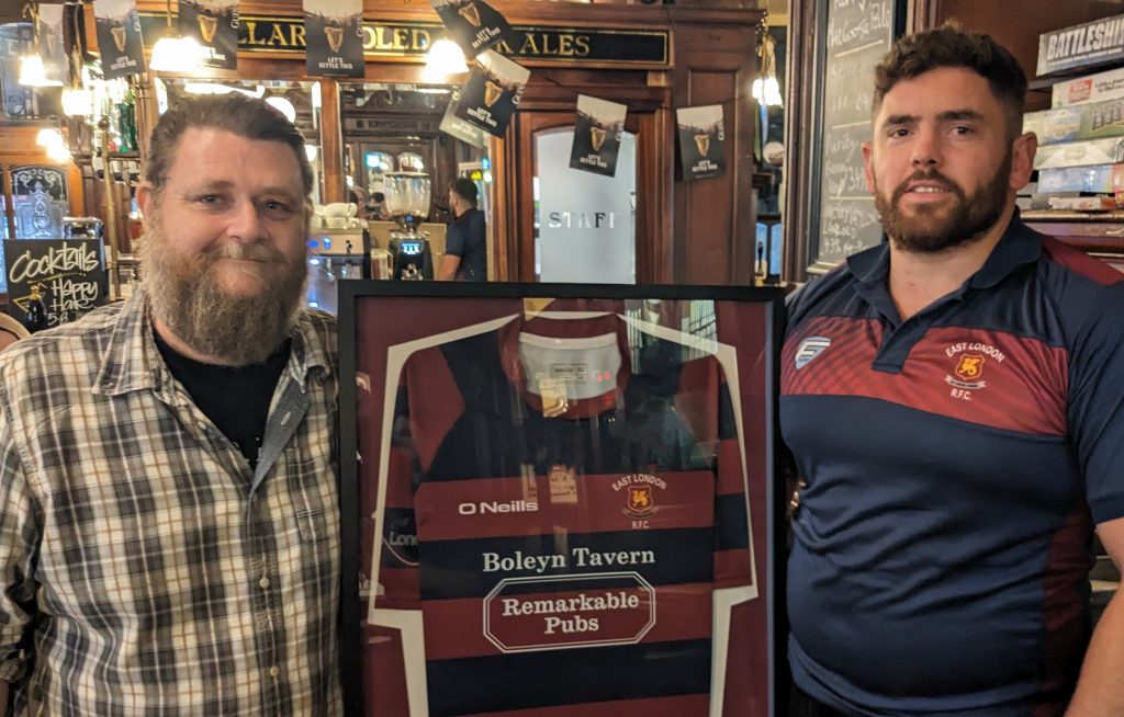 Two people in a pub pose next to a rugby shirt