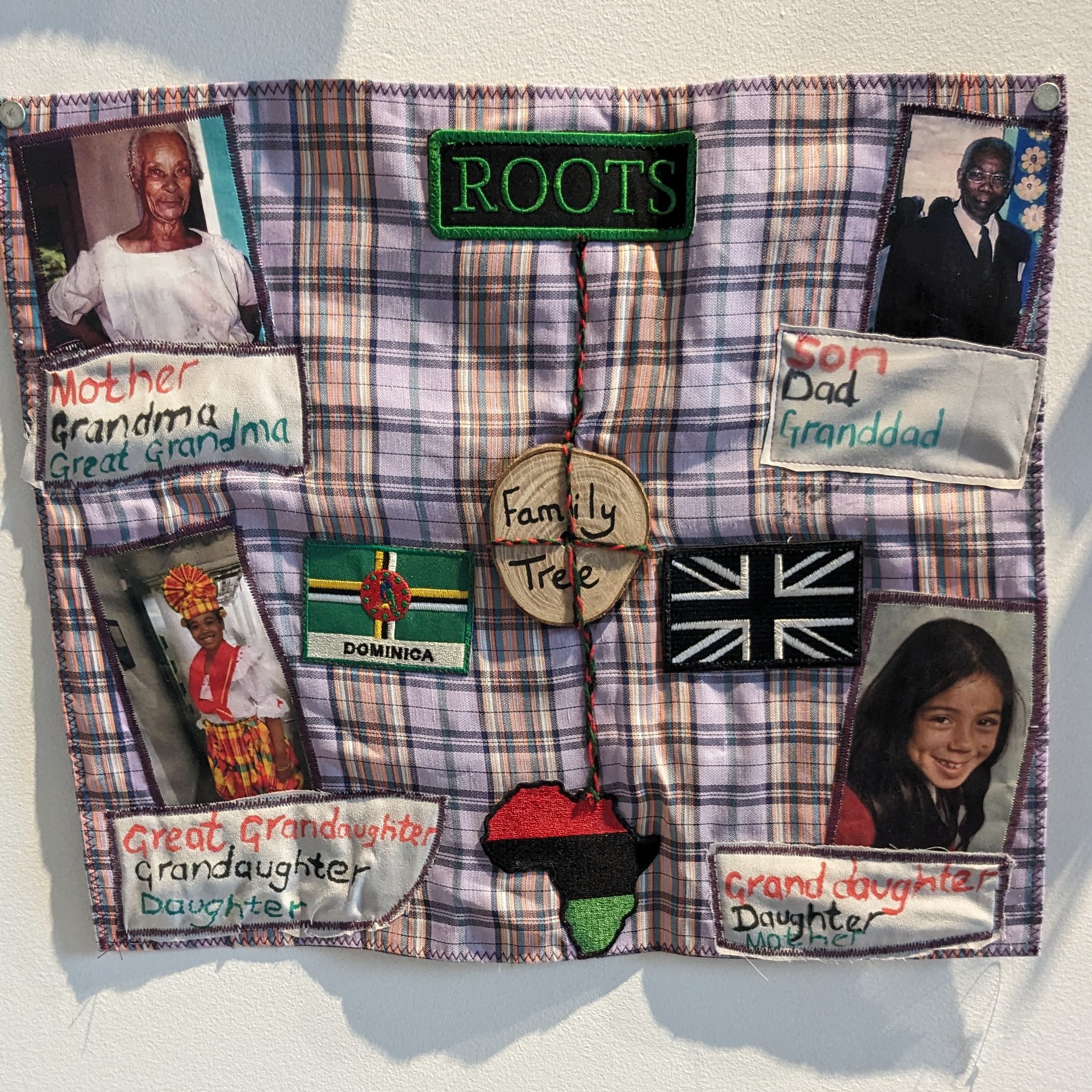 A piece of fabric with embroidered patches and photos of family hangs on a gallery wall