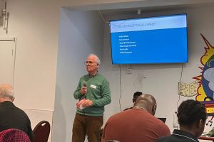 A person stands holding a microphone at the front f a meeting