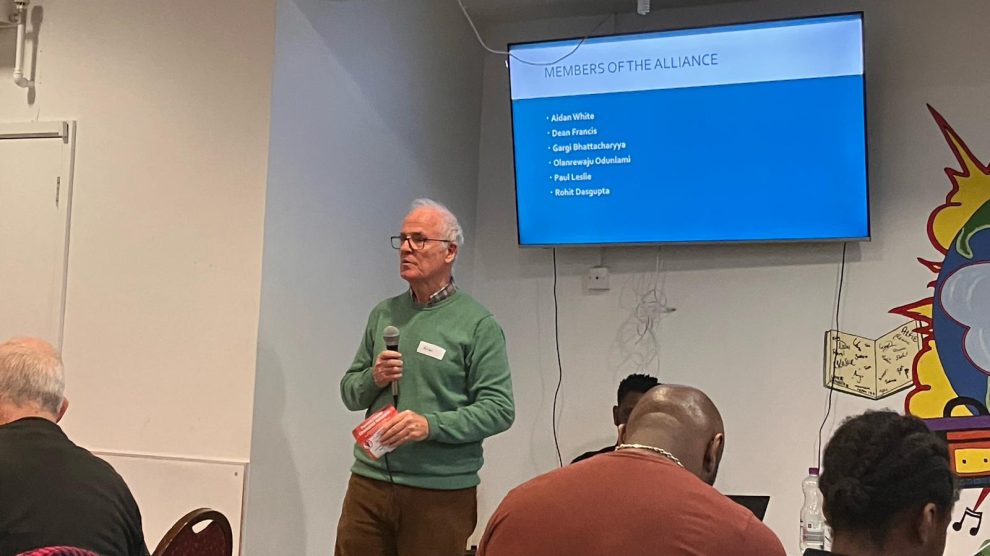 A person stands holding a microphone at the front f a meeting