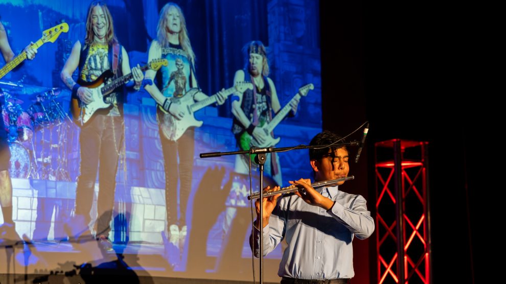 a person performs on a flute in front of a projection of people playing electric guitars