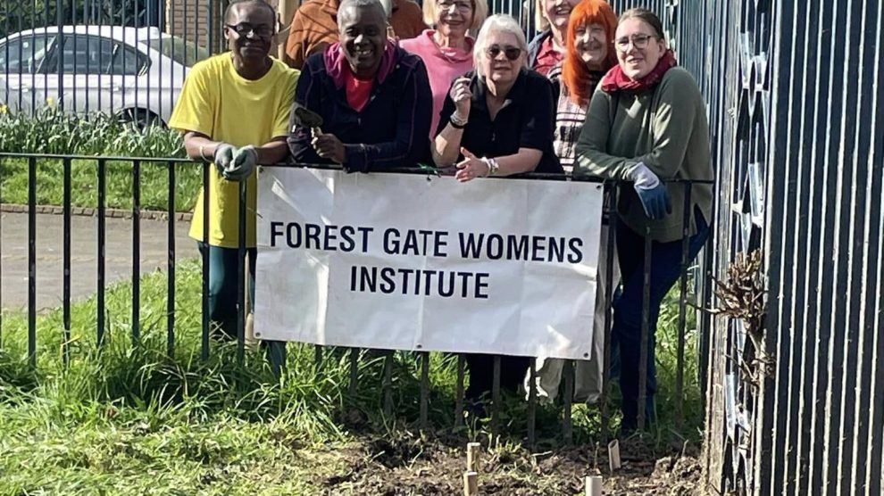A group of women stand behind a banner reading Forest Gate Women's Institute with newly planted saplings in the foreground