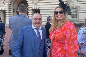 A smartly dressed man and woman stand in front of Buckingham Palace