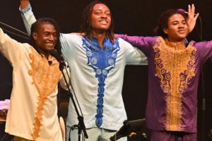 Three performers stand on stage arms aloft