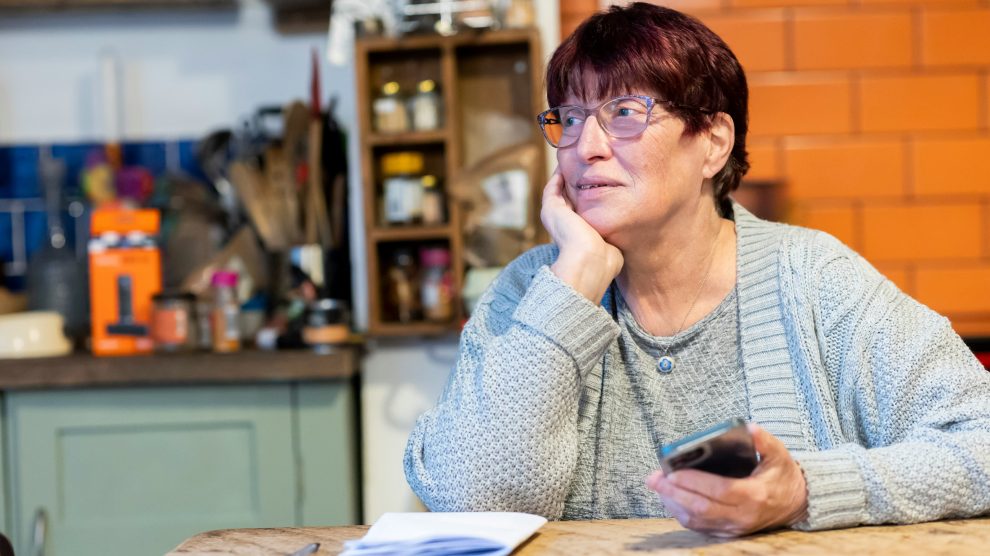 An older person sits at a kitchen table holding a mobile phone