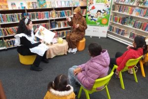 Three children in a library having a story read to them by a teacher or librarian