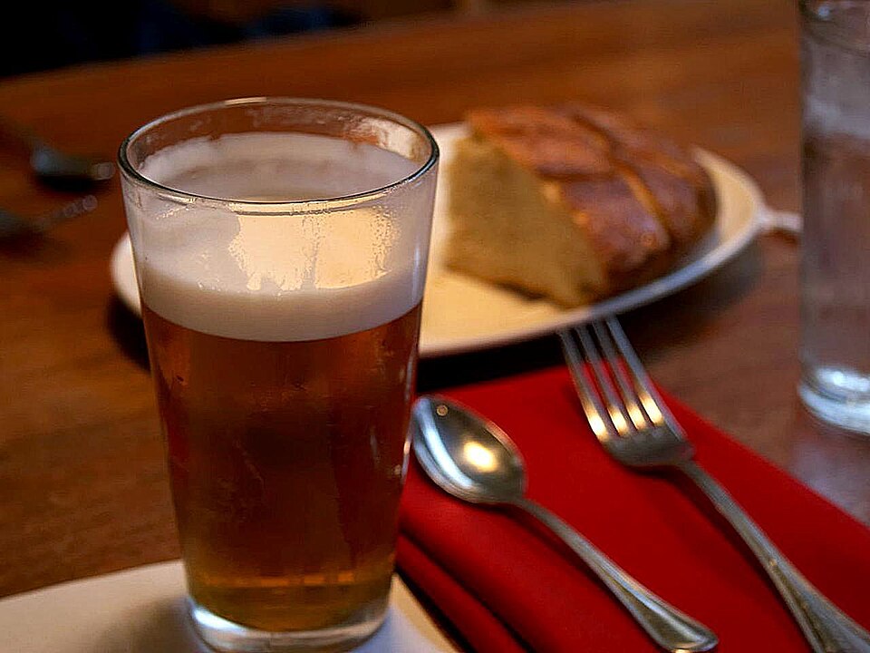 A pint of beer in the foreground with some bread on a plate in the background alongside some cutlery
